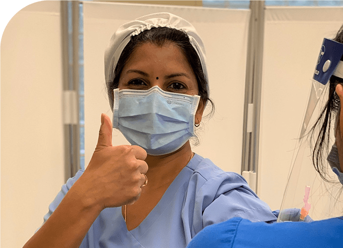 photo of a Priya Subramanian in a surgical mask and hat, giving the thumbs up sign