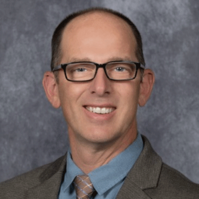 Aaron Sandquist - Texas Hospital Association Professional headshot of a smiling man wearing glasses, a blue shirt, brown blazer, and plaid tie against a gray backdrop.