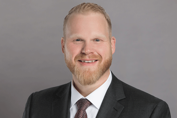 Professional headshot of a smiling man in a dark suit and white shirt against a gray backdrop.