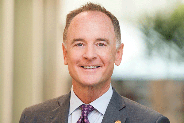Smiling middle-aged man in a gray suit and purple tie, posing for a professional headshot with a blurred office background.