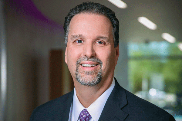 Professional headshot of a smiling man in a dark suit and purple tie in an office hallway.