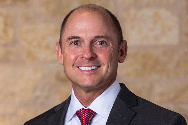 Smiling man in a dark suit and red tie, posed against a beige stone wall (professional headshot).