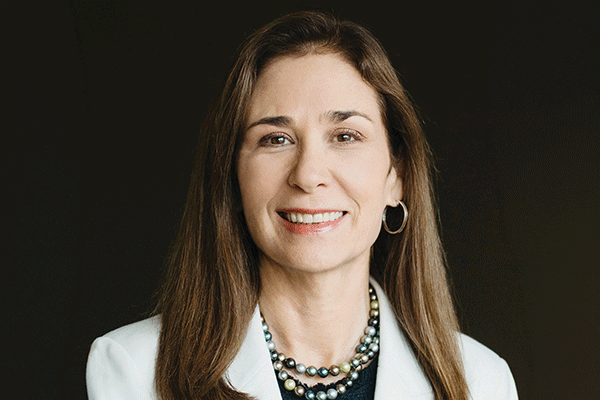 Professional head-and-shoulders portrait of a smiling woman with long brown hair, wearing a light blazer and layered pearl necklaces.