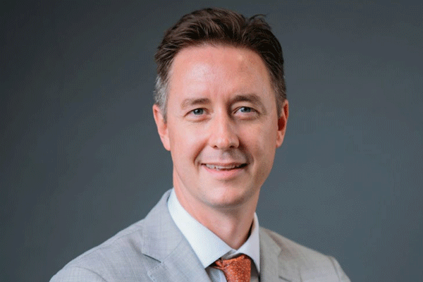 Head-and-shoulders portrait of a smiling man in a light gray suit and orange tie against a neutral gray background