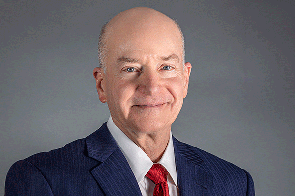 Professional headshot of a smiling bald man in a navy suit and red tie against a gray backdrop, looking at the camera.