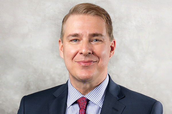 Head-and-shoulders portrait of a smiling man in a navy suit, checkered shirt, and red tie against a gray background