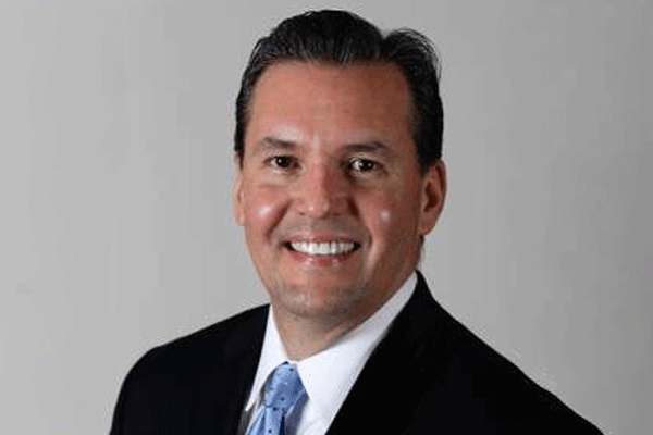 Professional headshot of a smiling man in a dark suit and blue tie against a neutral gray background.