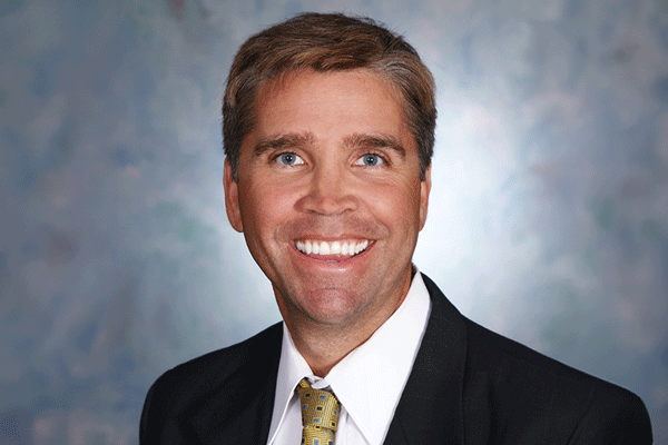 Professional headshot of a smiling man in a black suit and yellow patterned tie against a neutral blue-gray backdrop.