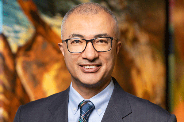 Professional head-and-shoulders portrait of a smiling man in a dark suit and glasses, with a blurred colorful background.
