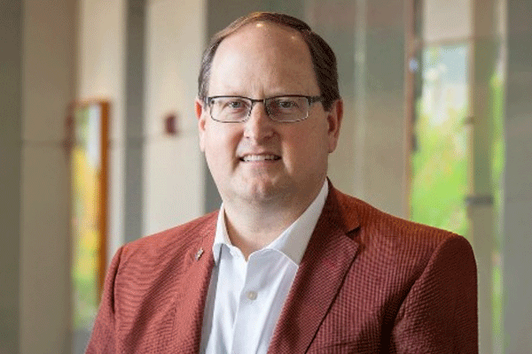 Professional headshot of a man with glasses wearing a white shirt and red blazer in a light office hallway.