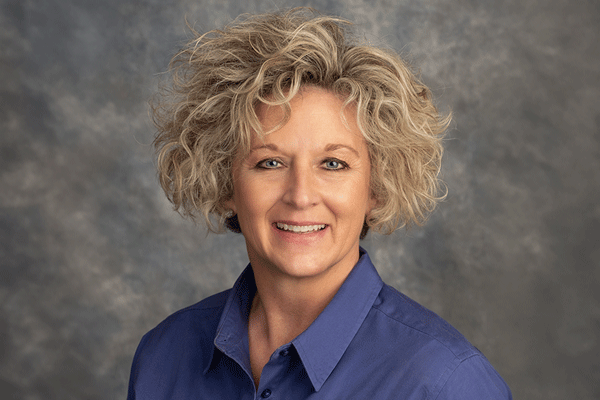 Smiling woman with short curly gray hair wearing a blue collared shirt, neutral studio backdrop behind her.