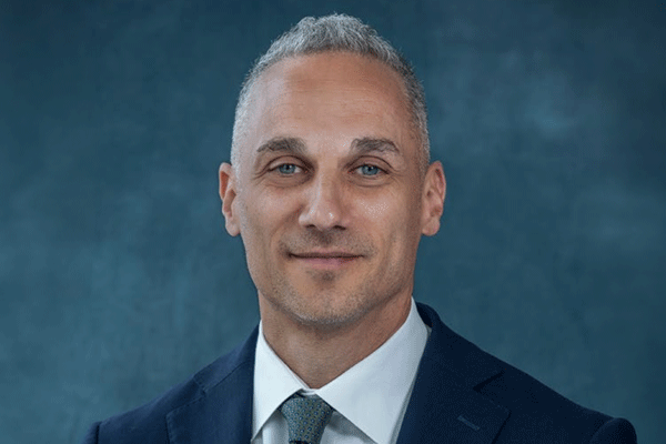 Professional headshot of a middle-aged man with short gray hair in a navy suit and teal tie against a blue backdrop.