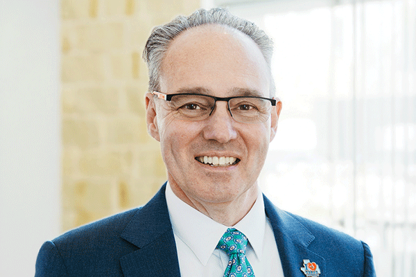 Smiling man in a navy suit and teal tie, wearing glasses—professional headshot in an office setting.