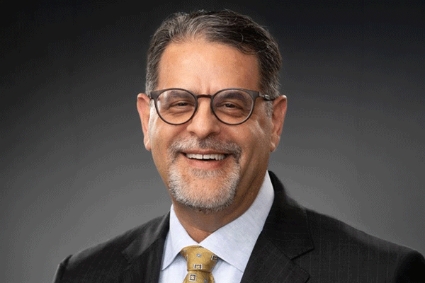 Smiling man in a dark suit and yellow tie, wearing glasses in a professional headshot.