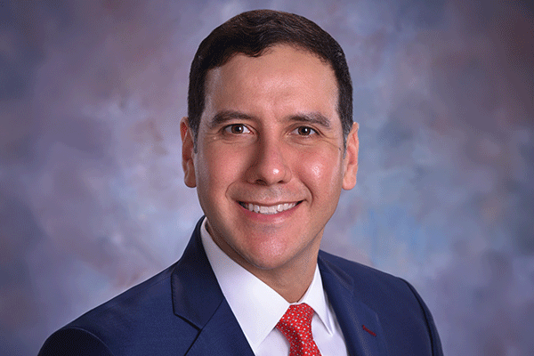 Professional headshot of a smiling man in a navy suit, white shirt, red tie, against a blue-gray backdrop.