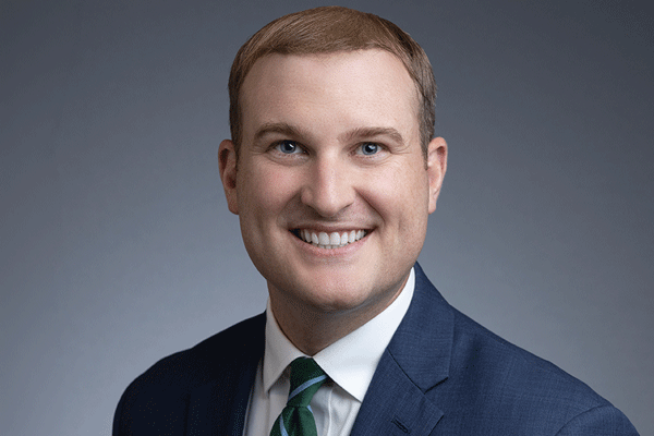 Professional headshot of a smiling man in a blue suit, white shirt, and green tie against a gray backdrop.