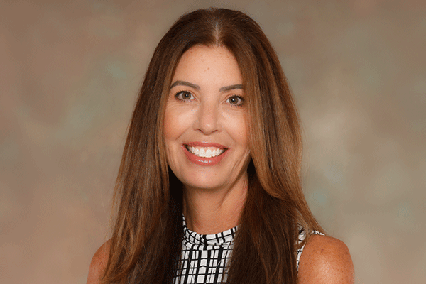 Portrait of a smiling woman with long brown hair wearing a black-and-white patterned sleeveless top against a neutral backdrop