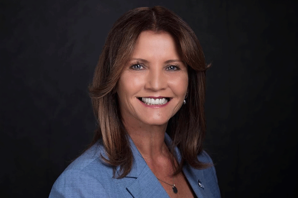 Professional portrait of a smiling woman with shoulder-length brown hair wearing a blue blazer against a dark background.