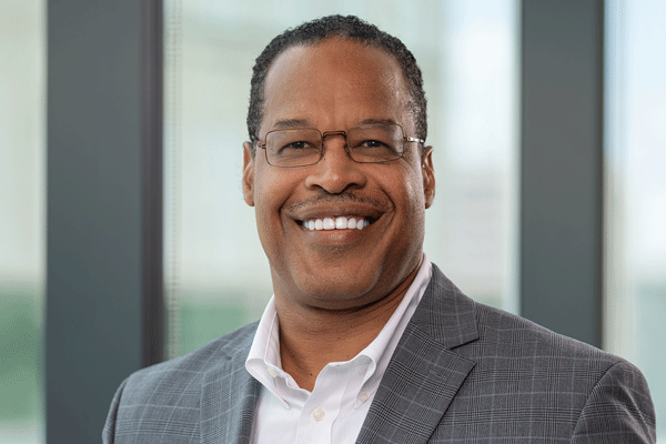 Smiling man in a gray blazer and white shirt, wearing glasses, in a modern office setting with large windows behind him on both sides.