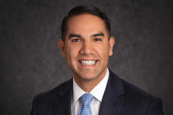 Professional headshot of a smiling man in a dark suit and blue tie against a neutral backdrop.