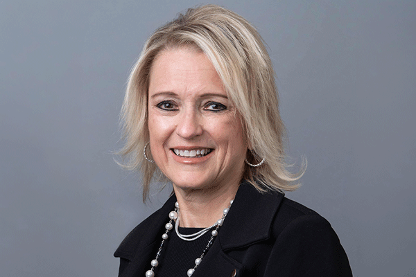 Professional woman with shoulder-length blonde hair, wearing a black blazer and pearl jewelry, smiling against a gray studio background.