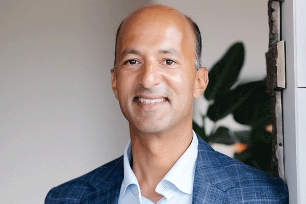 Professional portrait of a man in a blue blazer and white shirt smiling at the camera, indoors with plants in the background