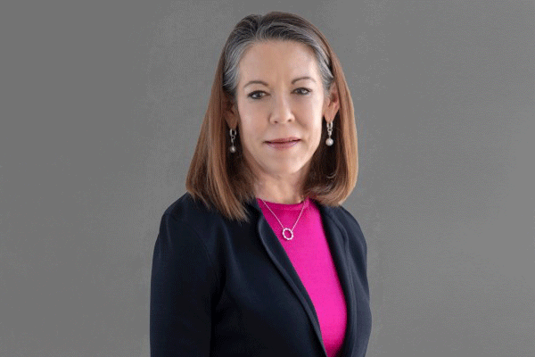 Professional head-and-shoulders portrait of a middle-aged woman in a navy blazer and pink blouse, against a gray background.