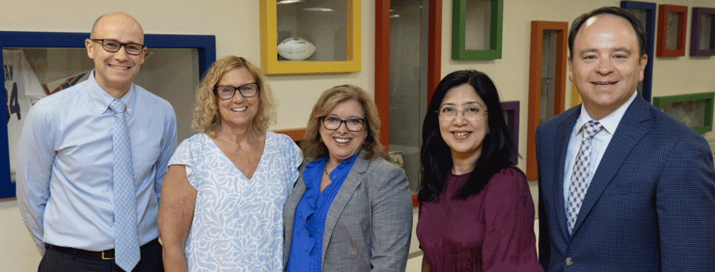 Five professionals posing together in an office, smiling for a group photo in business attire.