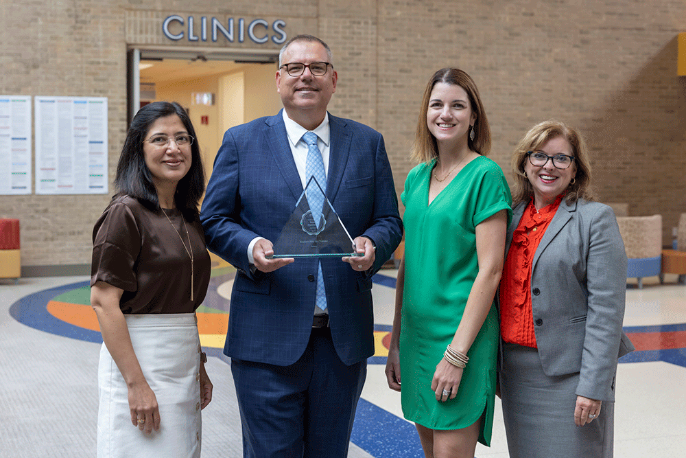 Four professionals stand in a clinic lobby; a man in a blue suit holds a glass award and smiles, flanked by two women and a companion in business attire.