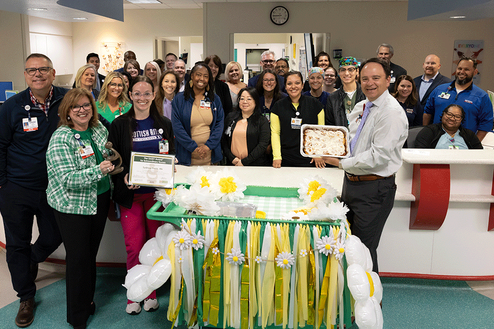 Group of healthcare workers posing in a hospital lobby with a decorated table and cake for a celebration, one person holding a certificate and another a tray of baked goods.