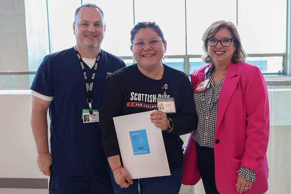 Three healthcare workers pose together in a bright hallway; center person holds a document or folder with a blue cover.