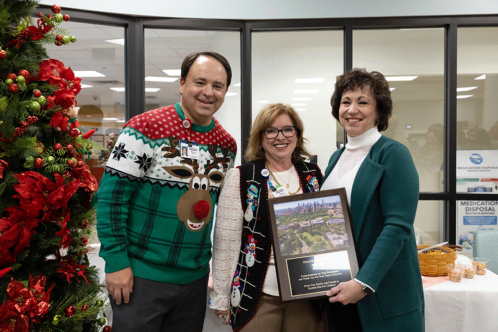 Three adults smile for a photo beside a Christmas tree; the woman in the teal coat holds a framed award with another woman and a man wearing a festive sweater nearby.
