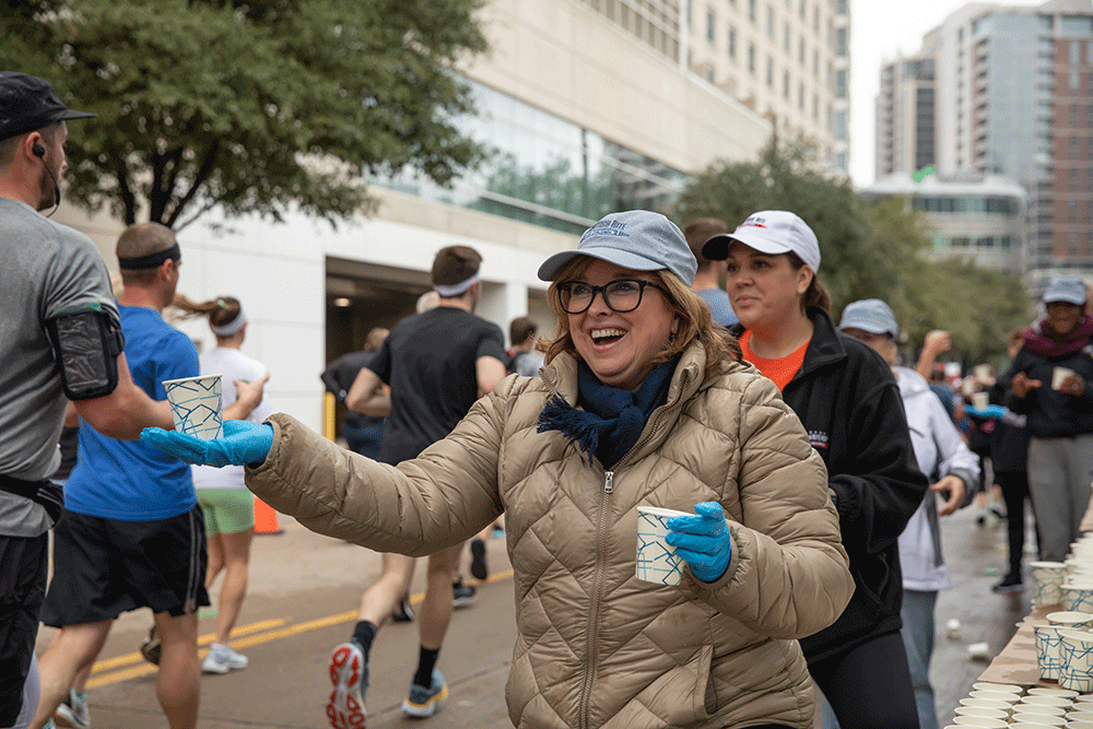 Smiling woman in a beige jacket and cap hands a cup to a runner at an urban race aid station. Several volunteers and runners in the background.