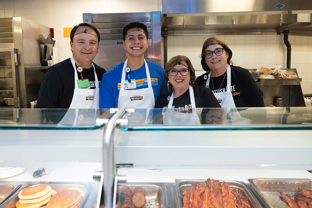 Four smiling coworkers behind a food counter wearing aprons in a busy kitchen setting.