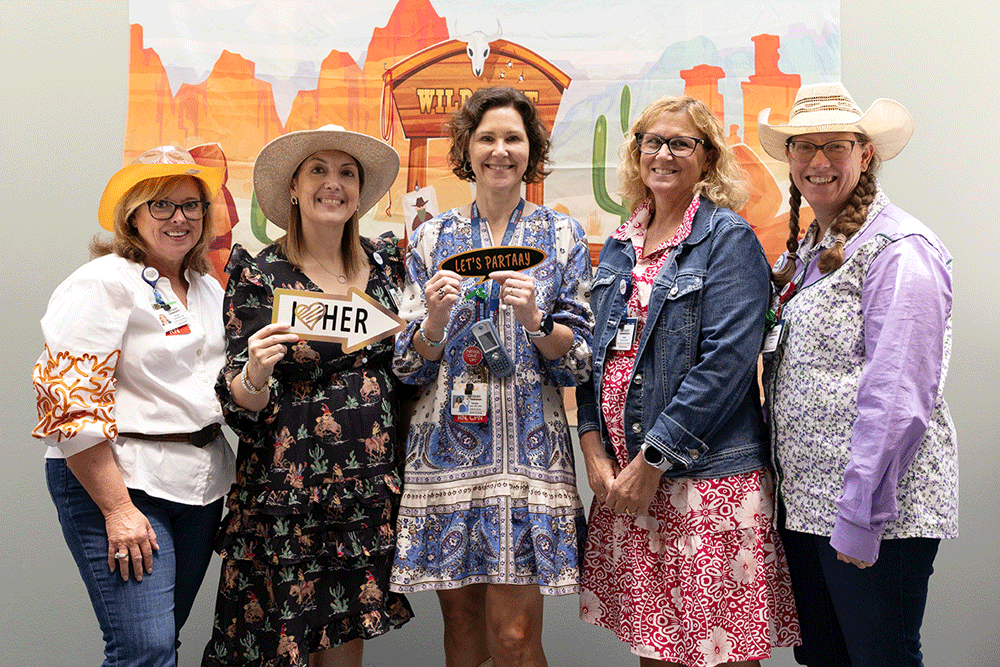 Five women in cowboy hats and colorful outfits pose together at a western-themed event, one holding a sign that says 'HER' and another 'LET'S PARTY' at center.