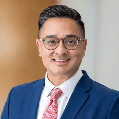 Joshua Pozos - Texas Hospital Association Portrait of a smiling man in a blue suit, white shirt, pink tie and glasses, in an office setting.