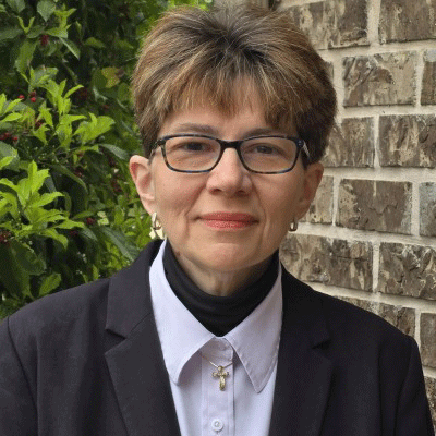 Melissa Winans - Texas Hospital Association Portrait of a woman with short light brown hair and glasses, wearing a dark blazer and white shirt with a cross necklace, standing beside a brick wall.