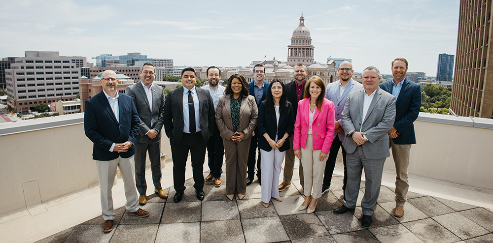 Twelve professionals posing on a rooftop with a city skyline and domed capitol building in the background.