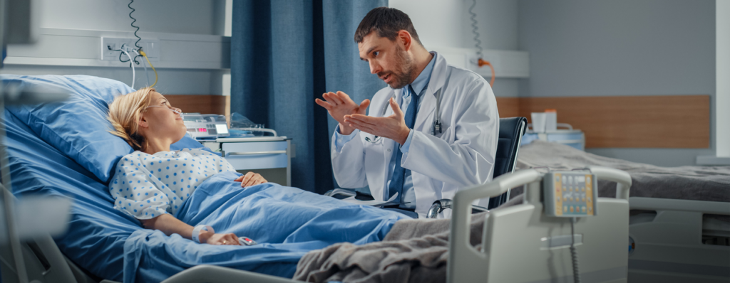 Patient Safety Organization - Texas Hospital Association Doctor in a white coat explains something to a patient lying in a hospital bed with medical equipment nearby.