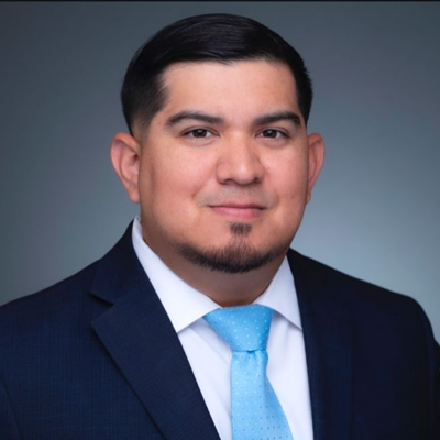 Robert Omar - Texas Hospital Association Professional headshot of a man in a dark navy suit, white shirt, light blue tie, smiling, against a blue-gray gradient background.