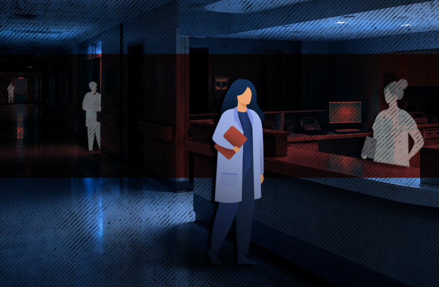 Healthcare worker in a blue coat holding a folder, standing in a dim hospital hallway.
