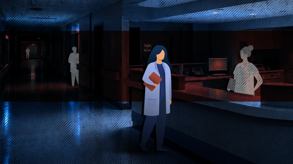 Healthcare worker in a blue coat holding a folder, standing in a dim hospital hallway.