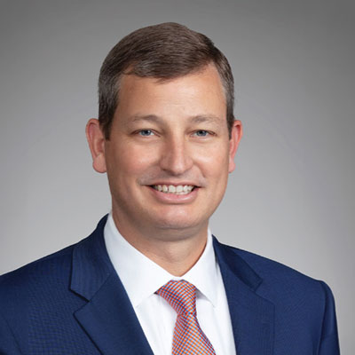 Brian Dean - Texas Hospital Association Professional headshot: smiling man in a navy suit with an orange tie against a gray backdrop.
