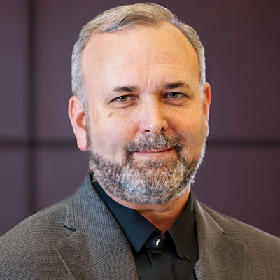 Terry-Scoggin-2026 - Texas Hospital Association Portrait of a middle-aged man with gray hair and beard, wearing a dark shirt and gray blazer.