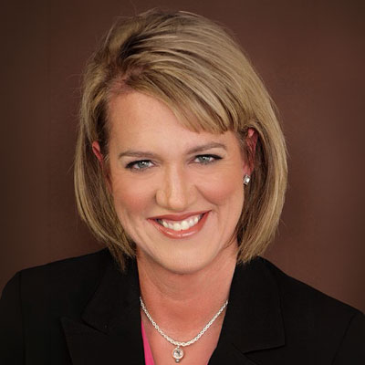 Wendi De Leon - Texas Hospital Association Professional headshot of a smiling blonde woman in a black blazer and pink blouse against a brown backdrop, wearing a silver necklace.