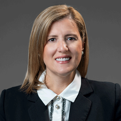 Edjola Xhaferi - Texas Hospital Association Professional headshot of a smiling woman in a black blazer and white blouse against a gray background.