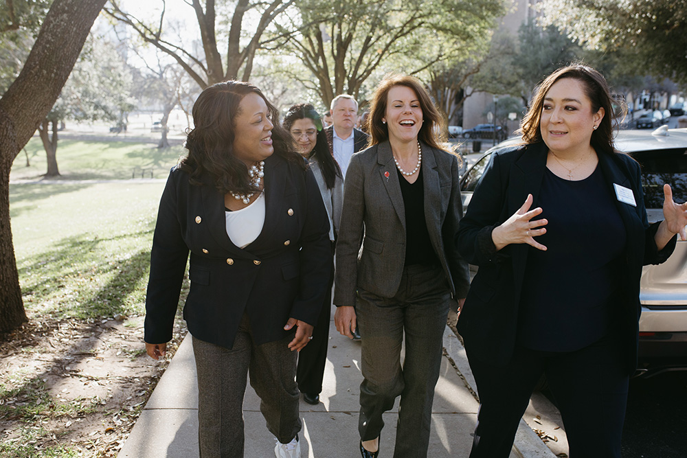 THA Fellows Class of 2026 on the way to visit the Texas Capitol - Texas Hospital Association Group of professional women walking together on a sunlit sidewalk, smiling and chatting.