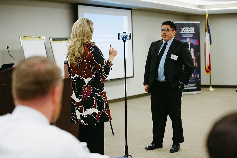 THA Fellows Class of 2026 in social media training - Texas Hospital Association Professional event scene: a man in a suit being interviewed by a woman in a patterned dress, filming with a smartphone on a tripod in front of a projector screen.