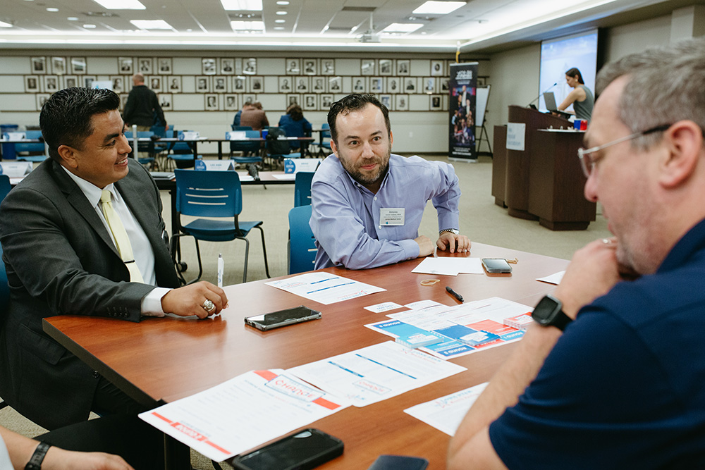 THA Fellows Class of 2026 practice making decisions - Texas Hospital Association Three men in a conference room sit around a table, discussing documents and notes strewn about the table.