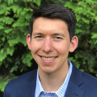 Luke Nakamoto - Texas Hospital Association Smiling man in a navy blazer and light blue check shirt posing outdoors with greenery behind him.
